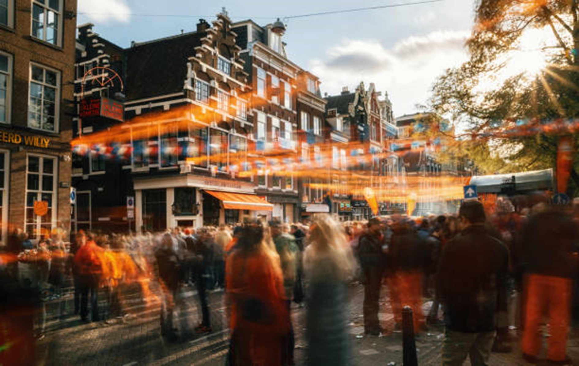 Een levendige foto van een kleine straat versierd in het thema van Koningsdag. Kleurrijke vlaggetjes wapperen in de wind terwijl mensen in oranje kleding langs de straat lopen. De straat wordt omzoomd door prachtige ouderwetse panden en de zon schijnt helder aan de blauwe hemel.